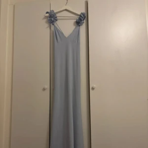 Ljusblå långklänning med volangdetaljer - Elegant ljusblå långklänning med smala axelband och v-ringning. Klänningen har fina volangdetaljer vid axelbanden och en knytning i ryggen för extra stil. Perfekt för bal! Aldrig använd! Bara testad! 