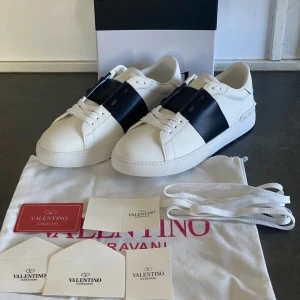 Valentino garavani skor - Snygga Valentino Garavani sneakers i vitt med en bred svart rem över snörningen. Skorna har en stilren design med en platt sula och diskreta detaljer. Perfekta för en trendig look.