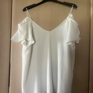 Vit ärmlös topp med volangdetaljer - Säljer en stilren vit topp med tunna axelband och volangdetaljer vid axlarna. Perfekt för en somrig look. Toppen har en luftig passform och är tillverkad i ett lätt material.