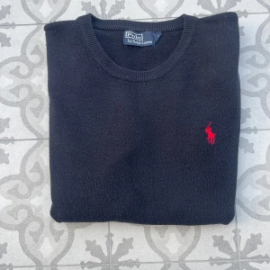 Stickad tröja från Ralph Lauren - Säljer en stilren svart  stickad tröja från Polo Ralph Lauren med det ikoniska röda logotypbroderiet på bröstet. Tröjan har långa ärmar och ribbade muddar vid ärmslut och nederkant. Perfekt för en klassisk look. Den är i storlek L men passar lite mindre. Hör av dig vid det minsta lilla! 🙌🤝
