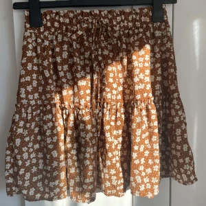Brun blommig kjol!! - Supersöt brun kortkjol med vita blommor och volangdetaljer. Kjolen har en bekväm elastisk midja och knytband framtill. Perfekt för en avslappnad och stilfull look för sommaren!🩷