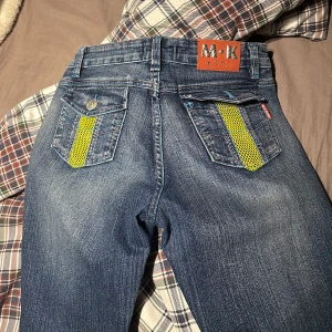 Blå jeans med gröna detaljer - Unika blå jeans med gröna nätliknande detaljer på fickorna. Byxorna har en bootcut-stil med fransade kanter och en låg midja. Perfekta för en trendig look med en twist. En knapp i bak på fickan är av och dom är lite smutsiga längst ner i bak