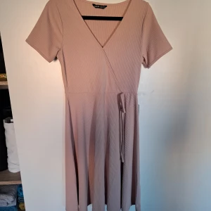 Beige ribbad klänning från New Look - Säljer en stilren beige klänning från New Look med ribbad struktur och v-ringad omlott-design. Klänningen har korta ärmar och ett knytband i midjan för en smickrande passform. Perfekt för både vardag och speciella tillfällen!