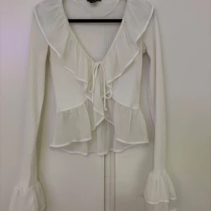 Vit ribbad blus med volangdetaljer i storlek XS - Säljer en vit ribbad blus med volangdetaljer och knyt framtill (man kan även knäppa under knuten). Använd några gånger. Skriv för fler bilder🤍