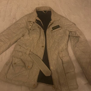 Beige quiltad jacka från Barbour - Säljer en snygg beige quiltad jacka från Barbour med bälte och dragkedja. Jackan har en klassisk look med Barbour International-märke på bröstet och praktiska fickor med knappar. Perfekt för en stilren och bekväm outfit.