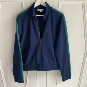 Blå wct-jacka från Carin Wester - Cool mörkblå jacka från Carin Wester med snygga gröna ränder längs ärmarna. Jackan har fickor och dragkedja framtill och är tillverkad i polyester och elastan. Storlek L. Axelbredd 45 cm, längd 58 cm. Knappt använd. 