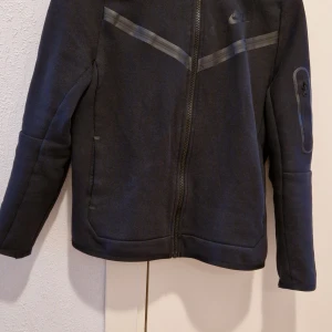 Svart fleecejacka från Nike  - Säljer en svart fleecejacka från Nike med dragkedja och huva. Jackan har en stilren design med diskreta detaljer och en ficka på ärmen. Perfekt för kyliga dagar och sportiga aktiviteter. Har använt 1 vecka. Den passar inte mig,därför säljer jag den populära Nike huddie. Storlek L,men det går använda XS/S osv.Köpte från Nike butiken i Emporia. Priset  kan diskuteras