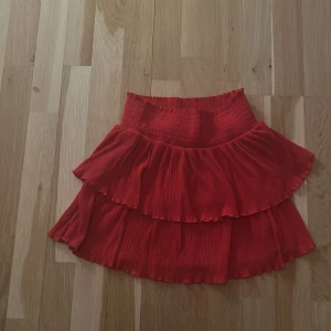 Röd volangkjol från Kappahl - Säljer en söt röd volangkjol från Kappahl. Kjolen har en elastisk midja och två lager volanger som ger en fin rörelse. Perfekt för att lägga till en färgklick i garderoben!