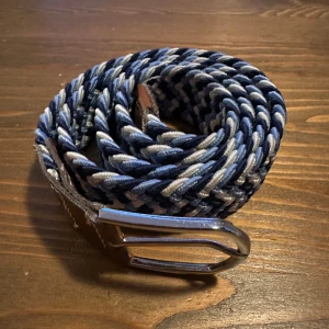 Flätat bälte i ljus blått, mörkblått och vitt - Snyggt flätat bälte ett stilrent silverfärgat spänne. Perfekt för att ge en extra touch till din outfit. Bältet har en unik design med flätat mönster som ger en modern känsla.