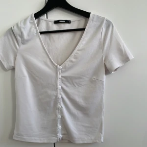 Vit kortärmad topp från Bik Bok - Säljer en vit kortärmad topp från Bik Bok med knappar framtill. Toppen har en v-ringad design och är perfekt för en avslappnad stil. Passar bra till jeans eller kjol.