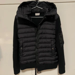 Moncler Svart Cardigan M - Säljer en snygg svart jacka från Moncler med quiltad design och stickade ärmar. Jackan har en dragkedja framtill och en huva för extra skydd. Perfekt för kyligare dagar!