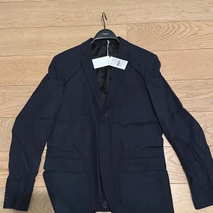 Mörkblå kavaj från Filippa K - Säljer en sprillans ny stilren mörkblå kavaj från Filippa K. Kavajen har en klassisk design med två knappar framtill och fickor med lock. Perfekt för en elegant look. Extra knappar ingår 