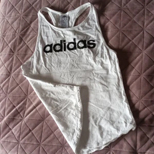 Vit cool topp från Adidas🦾💘 - Säljer en vit topp från Adidas med svart logga på framsidan. Perfekt för träning🤸‍♀️ eller en avslappnad dag. Den har en klassisk brottarrygg och är tillverkad i mjukt bomullsmaterial.💫🌸