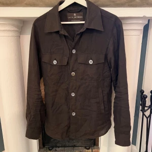 Brun jacka från Oscar Jacobson - Säljer en stilren brun jacka från Oscar Jacobson. Jackan har långa ärmar och knappar framtill samt två bröstfickor med knappar. Perfekt för en elegant och avslappnad look. Den är linne så perfekt för lite kyligare vår/sommar dagar. Nypris runt 4-5k
