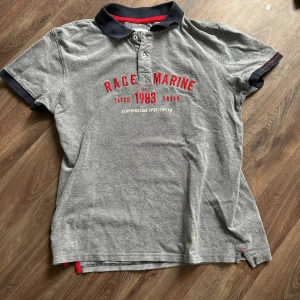 Grå pikétröja från Race Marine - Snygg grå pikétröja från Race Marine med röd text på bröstet. Tröjan har en klassisk krage och knappar framtill. Perfekt för en avslappnad stil.