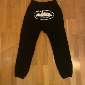 (Corteiz) Svarta mjukisbyxor med tryck - Säljer ett par svarta mjukisbyxor med ett coolt vitt tryck på baksidan. Byxorna har en avslappnad passform och resår i midjan och vid bensluten. Perfekta för en chill dag hemma eller en avslappnad utflykt.