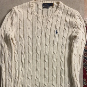 Vit stickad tröja från Polo Ralph Lauren - Säljer en stilren vit stickad tröja från Polo Ralph Lauren med klassiskt flätmönster. Tröjan har långa ärmar och en rund halsringning. Perfekt för kyligare dagar!
