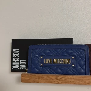 Blå quiltad plånbok från Love Moschino - Snygg blå plånbok från Love Moschino med quiltad design och guldfärgad logotyp på framsidan. Plånboken har en praktisk dragkedja och flera fack för kort och kontanter. ALDRIG ANVÄND! 