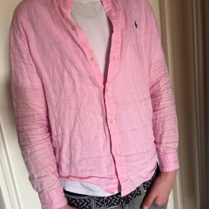 Rosa skjorta från Polo Ralph Lauren - Säljer en stilren rosa skjorta från Polo Ralph Lauren med klassisk krage och knappar framtill. Skjortan har en broderad logotyp i mörkblått på bröstet och är långärmad. Perfekt för en avslappnad men ändå uppklädd look. XL men sitter som en M
