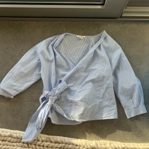 Blå- och vitrandig omlottopp från H&M - Säljer en snygg blå- och vitrandig omlottopp från H&M. Toppen har trekvartsärmar och ett knytband i midjan 💙