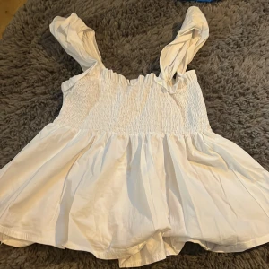 Vitt linne från Valentina - Charmigt vitt linne från Valentina med smockdetaljer och axelbandslös design. Perfekt för varma dagar med sin luftiga och bekväma passform. Säljer då jag inte andvänder men den är jättebra skick.