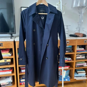 Mörkblå trenchcoat från Stockh LM - Snygg mörkblå trenchcoat från Stockh LM i storlek XS. Jackan har knappar framtill och ett bälte i midjan för en stilren look. Tillverkad i bomull med polyesterfoder, perfekt för vår och höst. En klassisk och tidlös design som passar till många tillfällen.