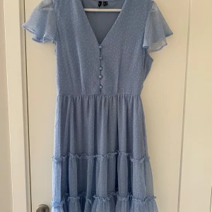 Ljusblå klänning med volanger - Charmig ljusblå klänning med prickigt mönster och volanger. Den har en v-ringad hals och knappar framtill. Perfekt för sommarens alla tillfällen.