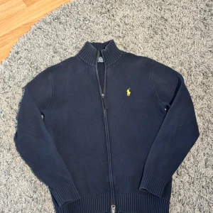 Mörkblå tröja med dragkedja från Ralph Lauren - Säljer en stilren mörkblå tröja från Ralph Lauren med dragkedja och ribbad krage. Den har ett broderat gult logotypmärke på bröstet och långa ärmar med ribbade muddar. Perfekt för en klassisk look.