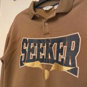 Brun pikétröja från H&M x Harry Potter - Säljer en brun pikétröja från H&M x Harry Potter-kollektionen. Tröjan har texten 'SEEKER' i blått och guld med en gyllene vinge under. Den har en klassisk krage och knappar framtill. Perfekt för fans av magiska äventyr! 🧙‍♂️
