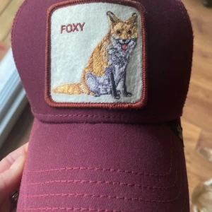Vinröd keps från Goorin Bros - Snygg vinröd keps från Goorin Bros med ett broderat rävmotiv och texten 'FOXY' på framsidan. Kepsen har ett nätmaterial på baksidan och justerbar passform.