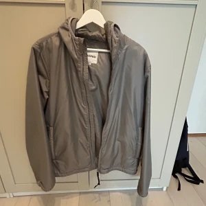 Grå vindjacka från Aspesi - Säljer en stilren grå vindjacka från Aspesi. Jackan har en praktisk huva och dragkedja framtill. Perfekt för blåsiga dagar och enkel att matcha med olika outfits. Köpt från Zalando för ca 4000, använt fåtal gånger 