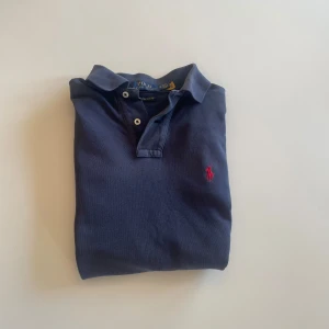 Blå pikétröja från Ralph Lauren - Säljer en stilren blå pikétröja från Ralph Lauren med klassisk krage och knappar. Tröjan har en broderad röd logotyp på bröstet och är i en slim fit-modell. Perfekt för en avslappnad men ändå snygg look.