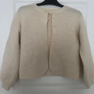 Beige stickad kofta med knappar - Mysig beige stickad kofta med rund hals och dekorativa knappar framtill. Perfekt för kyliga dagar med sin mjuka och bekväma passform. Koftan har långa ärmar och en fin struktur som ger en stilren look.