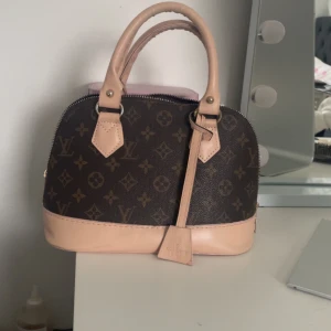 Brun handväska med monogram - Säljer en elegant brun handväska med det ikoniska monogrammönstret. Väskan har ljusa handtag och detaljer i skinn samt en dragkedja upptill. Perfekt för att ge en lyxig touch till din outfit. Kommer med dustbag !inte äkra!