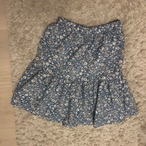 Blommig blå kjol - Söt blå kjol med blommigt mönster. Kjolen har en volangdetalj och är perfekt för sommarens varma dagar. Den har en bekväm passform och är ganska lätt att matcha med olika toppar.