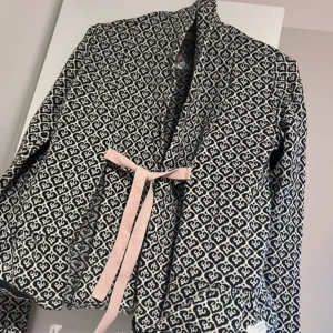 Svartvit mönstrad jacka med knytband - Säljer en unik svartvit mönstrad jacka från Desigual. Jackan har ett fint mönster och ett rosa knytband i midjan. Perfekt för att ge din outfit en extra touch!