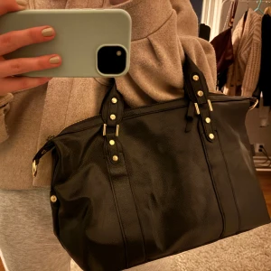 Svart handväska med dragkedja - Snygg svart handväska i skinn med dragkedja och dekorativa nitar. Perfekt för att bära dina dagliga nödvändigheter med stil. Väskan har praktiska handtag och en extra ficka med dragkedja på utsidan.