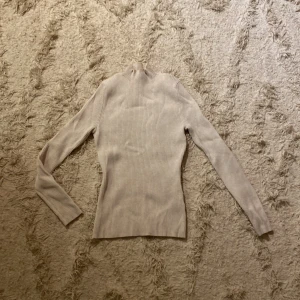 Beige ribbad polotröja - Säljer en stilren beige ribbad polotröja med långa ärmar. Perfekt för kyligare dagar och lätt att matcha med olika outfits. Tröjan har en hög krage som ger extra värme och stil.