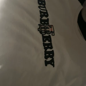 Vit t-shirt från Burberry - Säljer en stilren vit t-shirt från Burberry med deras ikoniska logga tryckt i svart och rött på bröstet. Perfekt för en casual look med en touch av lyx. Passar bra till jeans eller shorts för en avslappnad stil.