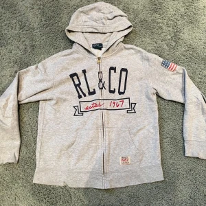 Grå zip hoodie från Polo Ralph Lauren - Säljer en grå hoodie från Polo Ralph Lauren med dragkedja och tryck framtill. Den har en amerikansk flagga på ärmen och texten 'RL CO est. 1967'. Perfekt för en avslappnad stil.