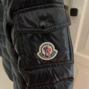 Moncler bady dunjacka - Snygg svart dunjacka från Moncler med huva och dragkedja. Passar bäst som höst-och vårjacka men också på vintern. Inte andvänd mycket alls pågrund av storleken är lite för liten. Pris kan diskuteras vid snabb affär. Kvitto finns. 