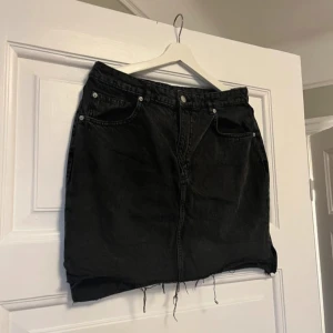 Svart jeanskjol - Snygg svart jeanskjol med klassisk femficksdesign och rå kant. Perfekt för en avslappnad stil. Kjolen har en knapp och dragkedja framtill. Storlek 42 men skulle säga mer 40.