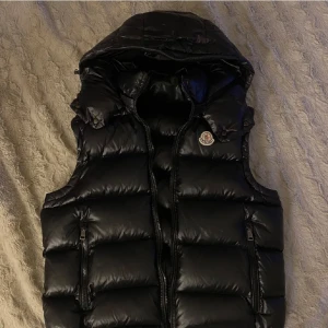 Svart dunväst från Moncler - Snygg svart dunväst från Moncler med dragkedja och huva. Perfekt för kyliga dagar och ger en stilren look. Västen har en klassisk design med Moncler-logga på bröstet. Skriv för mer bilder. Pris går att diskutera