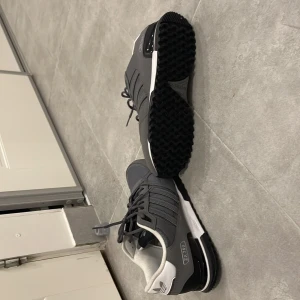 Grå sneakers från Adidas - Snygga grå sneakers från Adidas med klassiska tre ränder på sidan. Skorna har en vit sula och svart yttersula med bra grepp. De har snörning och en bekväm vadderad insida. Perfekta för en stilren look. Även bara använd i 1 tillfälligt 