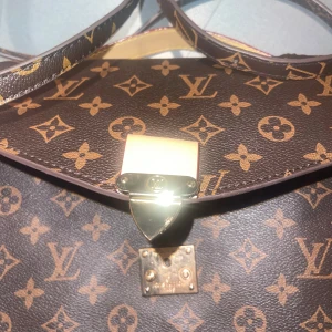 Brun monogram axelväska från Louis Vuitton - Säljer en elegant brun axelväska från Louis Vuitton med det ikoniska monogrammönstret. Väskan har en säker låsdetalj i guld och en praktisk dragkedja på insidan. Den röda insidan ger en lyxig känsla. Perfekt för att bära dina nödvändigheter med stil.