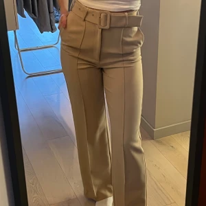 Beiga kostymbyxor med bälte från Ivyrevel - Snygga beiga kostymbyxor från Ivyrevel med hög midja och tillhörande bälte. Byxorna har raka ben, pressveck och en stilren look som passar perfekt till många olika outfits.