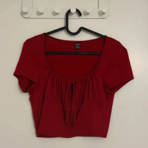 Röd croppad topp med knyt från SHEIN - Trendig röd croppad topp från SHEIN med korta ärmar och snygg knytning framtill. Toppen har en rundad urringning och lätt rynkade detaljer vid bysten. Perfekt för att lyfta din outfit med en pop av färg.