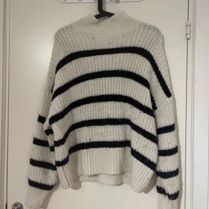 Vit och svart randig stickad tröja - Säljer en chunky stickad tröja i vitt med svarta horisontella ränder. Tröjan har hög krage och långa ärmar med breda muddar. Perfekt för kyligare dagar och ger en avslappnad, trendig look. Lite luddig är den lite överallt