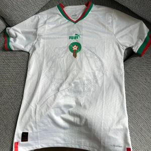 Marocko hemmatröja 2022 - Marockos landslagströja för hemmamatcher under 2022. Klassisk röd färg med gröna detaljer vid ärmar och krage. Puma-logga på bröstet samt landslagets emblem. Tunt och andningsbart material som gör den skön att bära i alla väder. Ett måste för fans av Marockos landslag