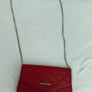 Röd quiltad axelväska från Love Moschino - Snygg röd axelväska från Love Moschino med quiltat mönster och guld kedjerem. Perfekt för att lyfta vilken outfit som helst med en pop av färg. Väskan har en elegant och stilren design med logotyp framtill.